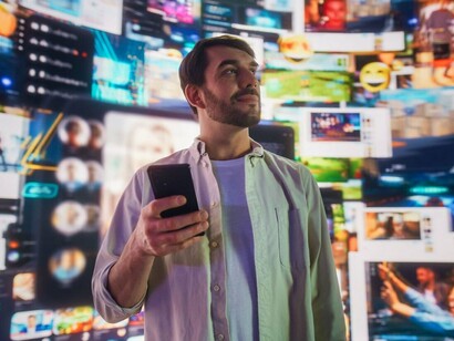 Com o smartphone na mão, um homem observa o ambiente repleto de imagens, vídeos e interfaces digitais ao seu redor. Em meio a tantos estímulos, encontrar aquilo que realmente desperta nosso interesse também se tornou uma tarefa dos algoritmos