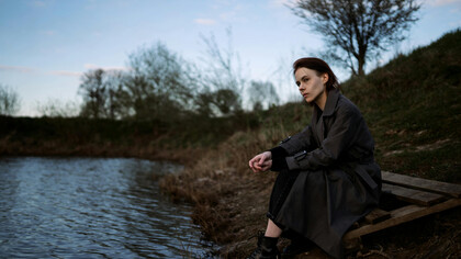 A young woman in a trenchcoat, contemplative, sitting at the shore of a small lake