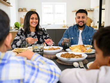 A three-generation Pakistani family shares a meal while a woman prepares food for the gathering