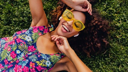 Central American woman enjoying summer vibes in a colorful hipster outfit, lying on grass and listening to music with wireless earphones