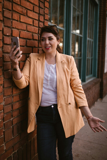 A woman in a beige coat and black pants stands beside a building at night, talking on the phone in a long-distance call