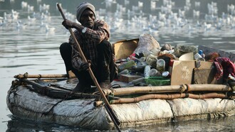 An adult man rows a makeshift raft filled with collected waste along a river, drawing attention to environmental pollution and cleanup efforts
