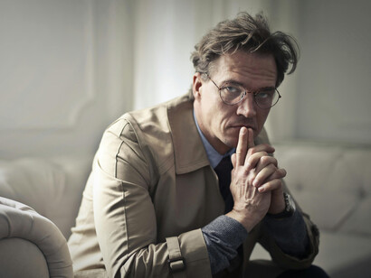 A man wearing a brown jacket sits quietly on a couch, appearing sad