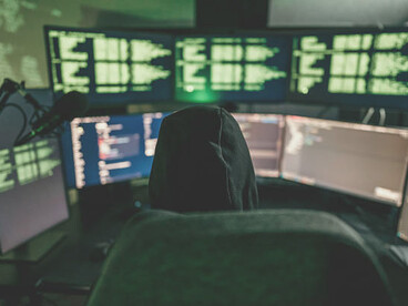 A dark-room setup with a hacker coding on multiple screens, illustrating cyber warfare and cyber threats
