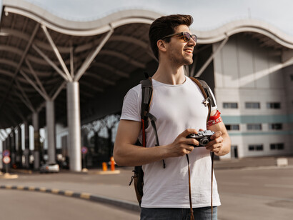A young solo traveler, dressed in a white T-shirt and sunglasses, smiling near an airport