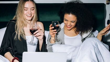 Two women with wine in hand looking at computer screen on bed and smiling