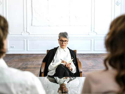A woman in a black suit, likely a psychologist, sits on a wooden chair while facilitating a couple’s therapy session