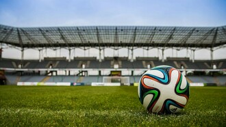 A lone soccer ball waits on the pitch, with an empty stadium looming in the background