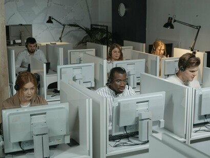 Uma equipe de suporte ao cliente trabalha em conjunto, conectada por computadores e fones de ouvido. São profissionais que integram uma estrutura indispensável ao funcionamento de muitos serviços, embora seu trabalho frequentemente permaneça invisível