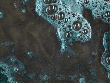 Foamy, chemical-laced water spreads across a dark surface, suggesting pollution beneath an abstract, almost organic pattern