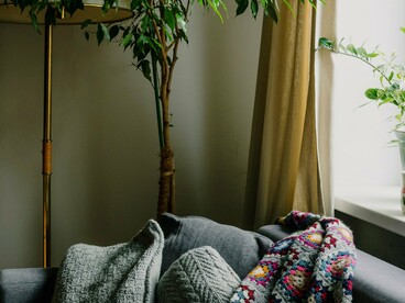 Um sofá, uma manta colorida e plantas ocupam uma sala iluminada, compondo um ambiente de aspecto acolhedor e vivido. A casa aparece aqui menos como cenário e mais como espaço de conforto, memória e experiências cotidianas