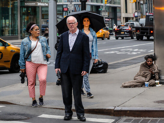 Busy city intersection in the USA highlighting social inequality, as pedestrians move through an urban street while a homeless woman sits unnoticed on the sidewalk
