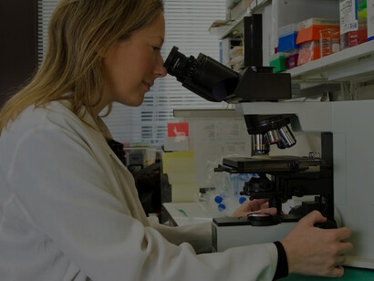 In a laboratory setting, a female scientist wearing a white lab coat sits on a black office rolling chair, attentively peering through a microscope