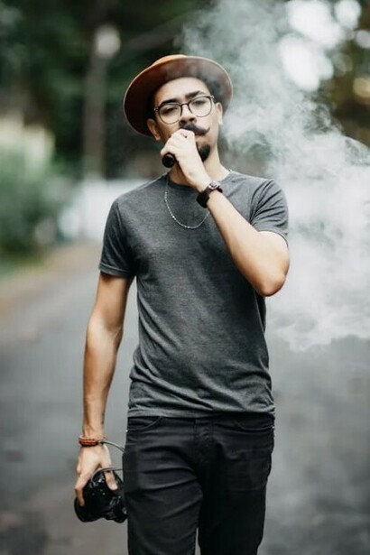 A man wearing a hat, glasses, and a mustache, dressed in a basic t-shirt and sneakers, striking a casual pose