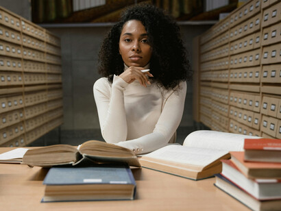 A woman researching at a library table surrounded by books