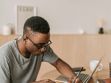 A man in a gray shirt uses a laptop for academic writing