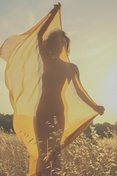 In the midst of a sprawling field, a woman stands with poise, her hand gently grasping a billowing scarf. The image captures a moment of natural elegance and tranquility