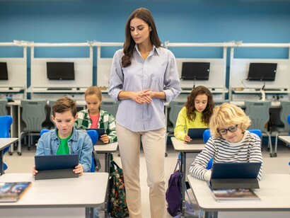 In un'aula, una maestra si trova in piedi, al centro, e guarda i suoi studenti mentre lavorano sui tablet
