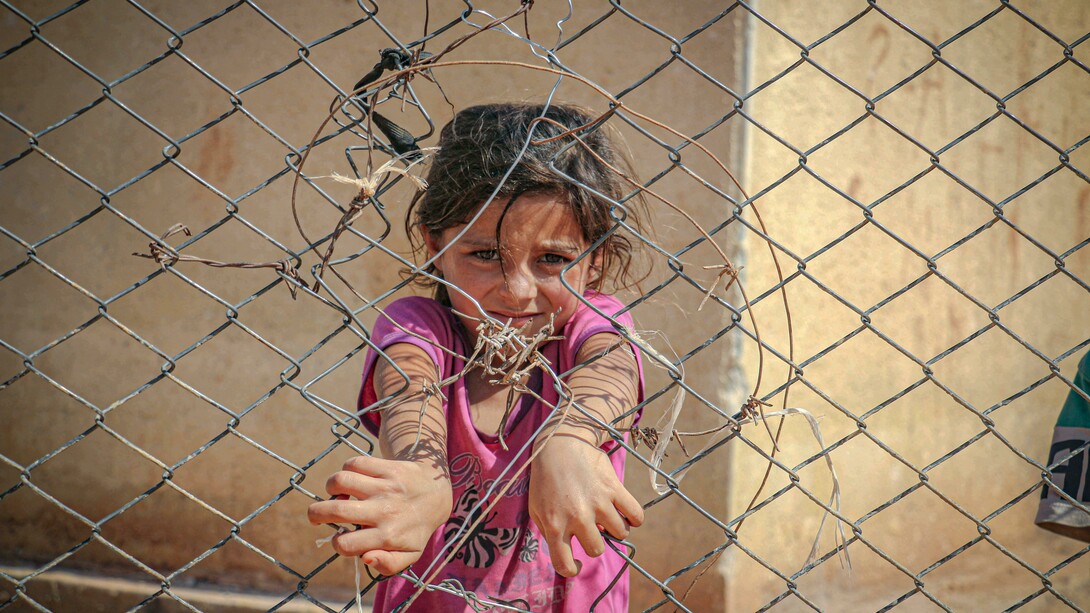 A young girl grips a chain-link fence in Idlib, Idlib Governorate, Syria