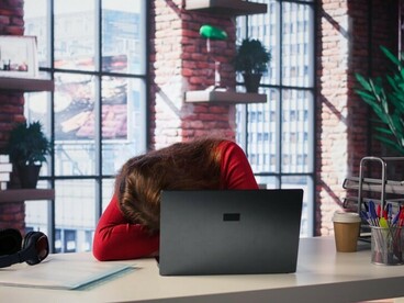 Exhausted and burned out, a woman sits in her loft, staring at her laptop screen and yawning