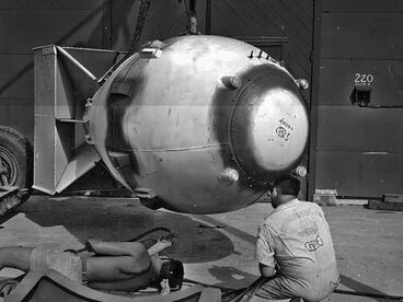 Homens trabalham ao lado da Fat Man durante os preparativos para seu emprego, enquanto a estrutura da bomba ocupa o centro da imagem. A cena evidencia como a tecnologia nuclear transformou a capacidade humana de produzir destruição em uma questão de engenharia e logística