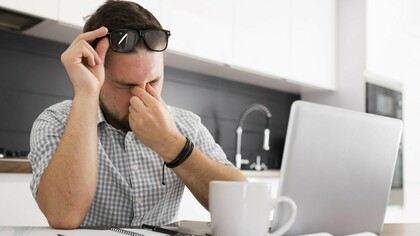 A man rubbing his eyes and looking overwhelmed while seated at a laptop,  indicating the physical and mental strain of occupational fatigue