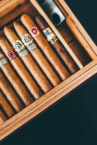A hand-made wooden cigar box, open to reveal a neatly arranged row of cigars with varied bands, alongside a silver cigar cutter