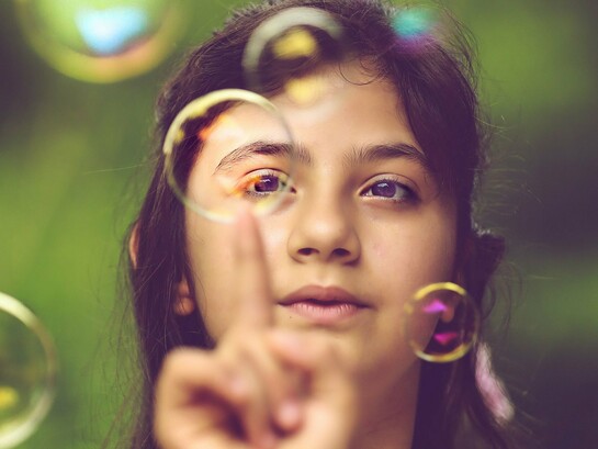 Com o olhar atento, uma menina toca com a ponta do dedo uma bolha de sabão, como se quisesse descobrir o que acontece ao entrar em contato com ela. Frágil e efêmera, a esfera desperta a curiosidade e transforma uma simples brincadeira em uma experiência de exploração do mundo. Na infância, gestos como esse revelam a disposição para observar, experimentar e descobrir, construindo conhecimentos a partir da interação com o ambiente. Os estudos de Jean Piaget ajudam a compreender como essas pequenas experiências participam do desenvolvimento cognitivo, mostrando que aprender também é agir, investigar e atribuir sentido àquilo que nos cerca