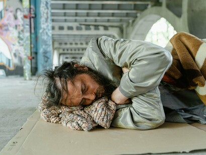 Um homem em situação de rua dorme sobre um pedaço de papelão sob um viaduto. A cena evidencia a precariedade das condições de descanso e proteção de quem não dispõe de moradia