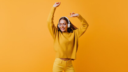 A carefree brunette woman in stylish yellow pants joyfully dancing to a good song, against a backdrop of an inviting orange wall, exuding warmth and happiness