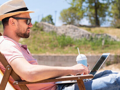 Man working on a laptop from a beach chair with a drink, showcasing a flexible summer work environment and work-life balance