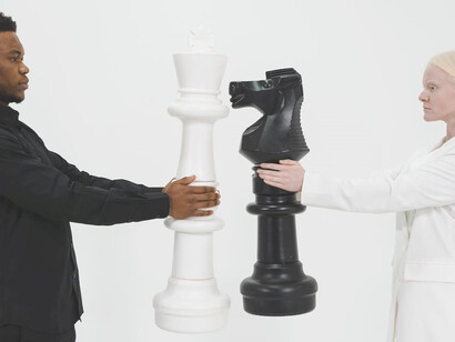 In a striking contrast, a woman dons an elegant white suit while a man exudes sophistication in a black suit. She cradles the black chess knight, and he holds the white rook, embodying a game of strategy and elegance