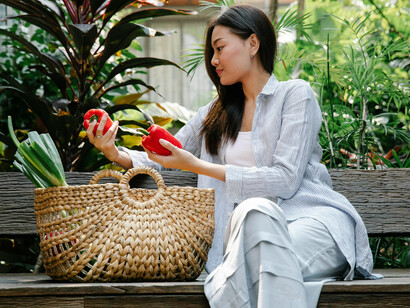 An Asian woman sits on a bench, gently gathering fresh vegetables into a wicker basket, reflecting a calm and simple way of life