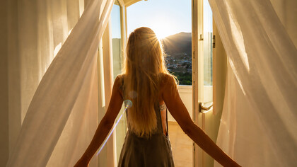 Golden light spills through the open doorway as a girl welcomes the morning, caught between quiet solitude and the promise of a new day