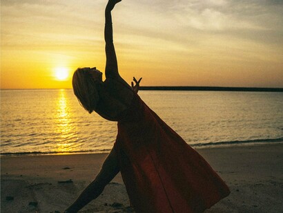 Mulher dança na praia. Coloque uma mão no coração, outra no ventre. Respire. E pergunte a si mesma: Como posso transformar essa resistência em reverência? O que posso honrar neste exato momento, mesmo que ainda esteja em construção?