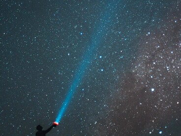 Uma silhueta aponta uma lanterna para o céu coberto de estrelas durante a noite. Tal como os escritores citados no texto, o gesto parece procurar respostas na vastidão infinita