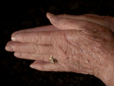 O enquadramento aproxima o olhar das mãos envelhecidas e de um anel dourado, destacando os detalhes da pele e do adorno. Mais do que um objeto, o anel pode evocar lembranças, afetos e experiências acumuladas ao longo da vida. O tempo transforma a aparência, mas o significado de uma existência não se mede pela juventude do corpo