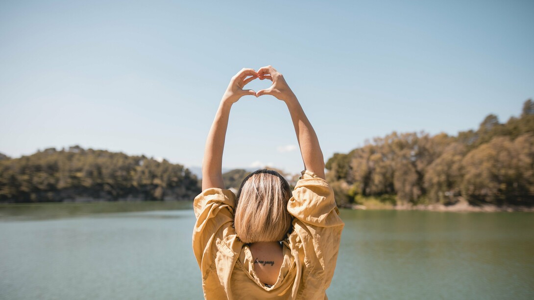 A woman  by a serene lake, her arms extended above her head, forming a heart shape with her hands against a clear blue sky, an image that embodies the balance between nature, self-care, and resilience