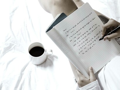 A woman on her bed with a cup of coffee writing in hour journal about things she doesn't understand but doesn't care to 