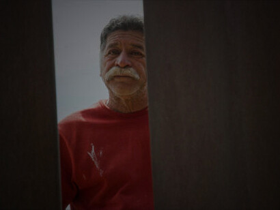 A Hispanic man in his sixties, sporting a mustache, gazes directly at the camera from the Mexican side of the steel-slat border wall separating Mexico and the United States
