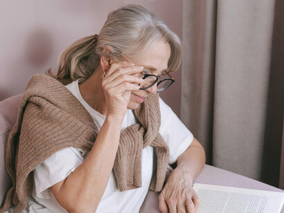 Elderly woman with glasses relaxing on a couch while reading a book at home