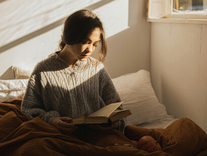 A young woman peacefully reading a book on her bed, as the early sunshine filters thorugh the windows