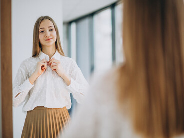 A young woman dressed casually gazes into the mirror, appearing confident