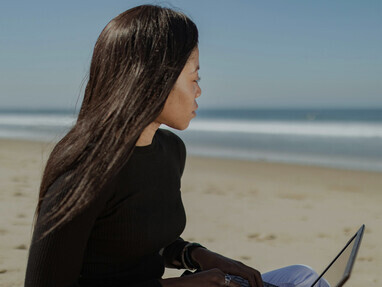 A woman in a black long-sleeve shirt sits by the beach, working on her laptop with the calm ocean as her backdrop