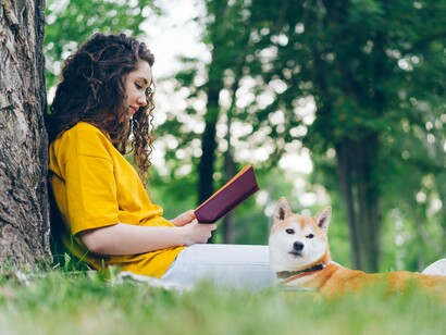 A woman seated on a blanket beneath a tree, absorbed in a book as her dog accompanies her, symbolising the grounding influence of simple, device-free moments