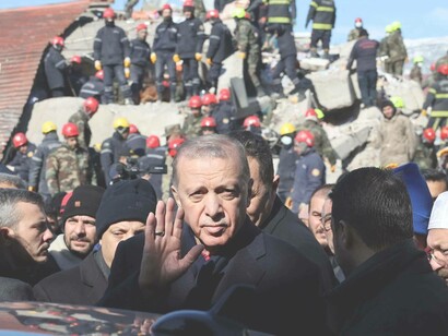 Turkish President Recep Tayyip Erdogan waves to the crowd as rescuers in the background deal with the collapsed buildings in Kahramanmaras, Turkey