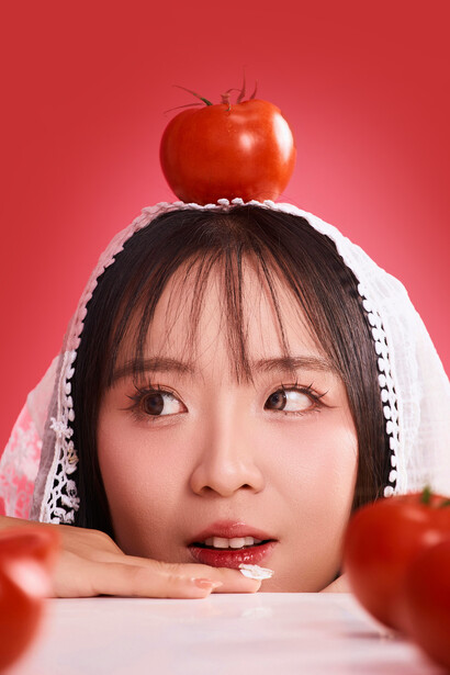 A playful, whimsical portrait of a woman with a tomato atop her head, photographed against a pink backdrop