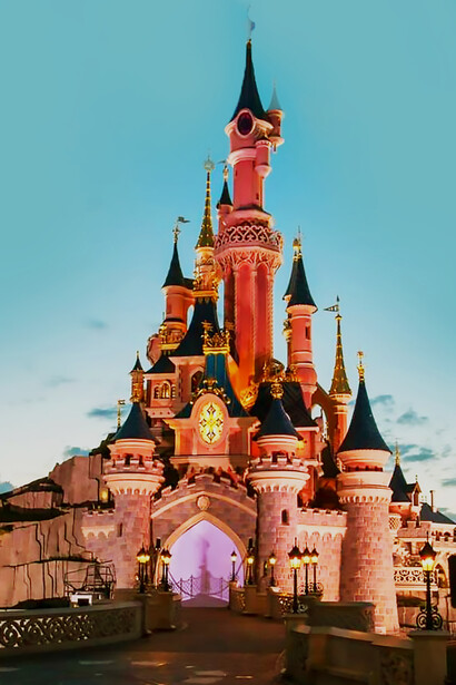 People stroll along the sidewalk next to a brown concrete building on a sunny day at Disneyland Paris, Marne-la-Vallée, France