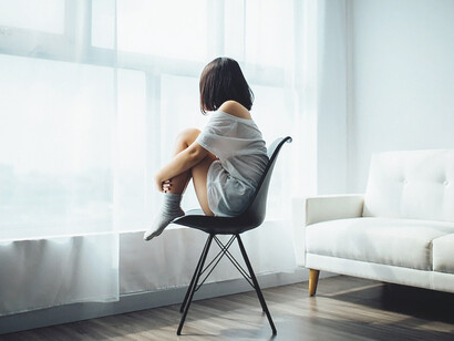 An elegant woman rests upon a black chair, framed by a glass-panel window adorned with billowing white curtains