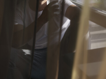 A contemplative man sits alone near a window, with soft natural light and curtain shadows creating an introspective atmosphere